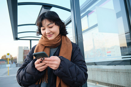 漂亮的日本女孩，学生穿着冬装站在公交车站，拿着智能手机，使用应用程序跟踪她的公共交通，叫车