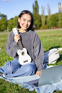 微笑的亚洲女孩学习如何在笔记本电脑上弹奏四弦琴，与音乐老师进行视频聊天，在公园的草地上与乐器坐在一起