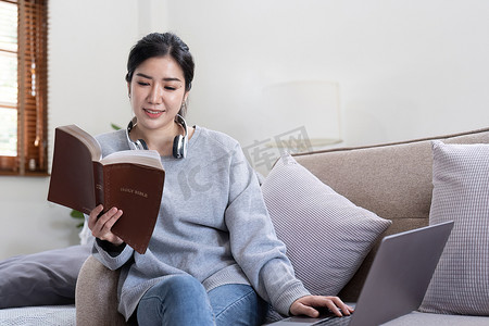 火爆进行中摄影照片_年轻漂亮的亚洲女学生坐在沙发上在家看书，同时使用笔记本电脑进行在线学习