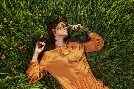 一个长着红色长发的平静女人躺在黄色花朵的绿色田野里，穿着橙色的连衣裙，闭着眼睛，双臂张开，享受着平静和休养