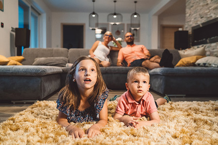 两个小孩躺在毛茸茸的地毯上看电影，父母坐在沙发上