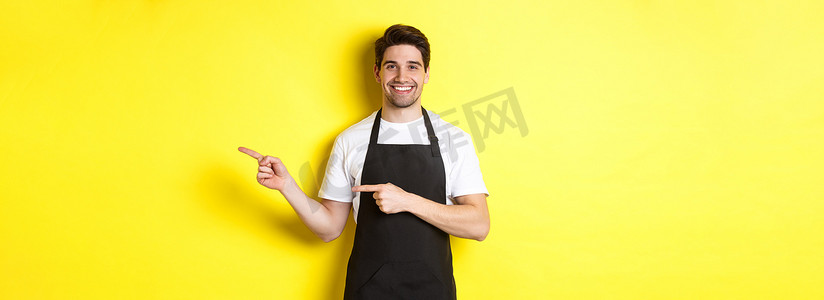 促销黄色背景摄影照片_友好的服务员用手指向右，展示您的标志或促销优惠，穿着黑色围裙制服，站在黄色背景上