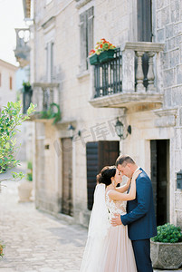 阳台人摄影照片_新郎在一座带阳台的石头建筑附近拥抱穿着白色连衣裙的新娘