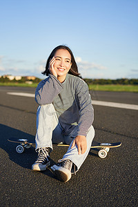 春天户外运动女摄影照片_亚洲女滑手的垂直镜头，坐在她的滑板上微笑，享受阳光明媚的日子，在户外空荡荡的路上在长板上巡航