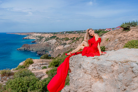 飘动摄影照片_一位身着红色丝绸连衣裙的女子坐在海边，背靠群山，她的裙子在风中摇曳。