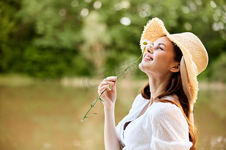 特写：一位戴着柳条帽、手拿洋甘菊的快乐女人在湖的背景上