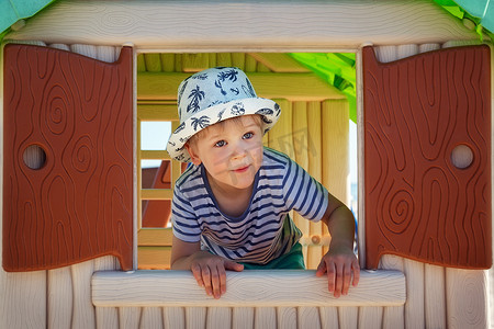 窗外操场摄影照片_一个穿着水手衫的可爱小男孩惊讶地看着玩具屋的窗外。