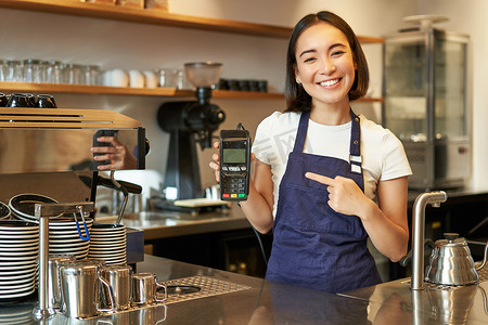 微笑的亚洲咖啡师女孩，穿着围裙，展示处理付款的信用卡机，建议非接触式付款，站在咖啡店