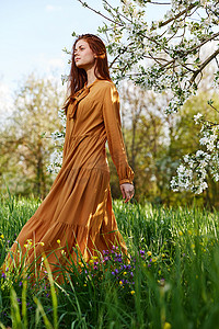 绿色树苗条摄影照片_一个苗条、甜美的女人穿着橙色长裙站在一棵开花的树旁高高的草丛中，看着别处，她的裙子在微风中展开