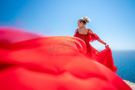 阳光明媚的衣服摄影照片_金发碧眼的长发在阳光明媚的海滨穿着红色飘逸的连衣裙，后视，丝绸面料在风中飘扬。