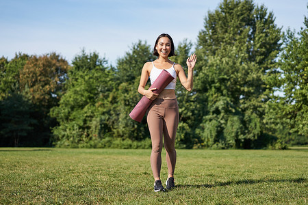 带橡胶垫的时尚女运动员打招呼，向你挥手，准备在草坪上的公园进行户外锻炼