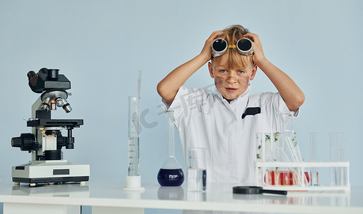 扮演了摄影照片_穿着外套的害怕的小男孩用设备在实验室里扮演科学家