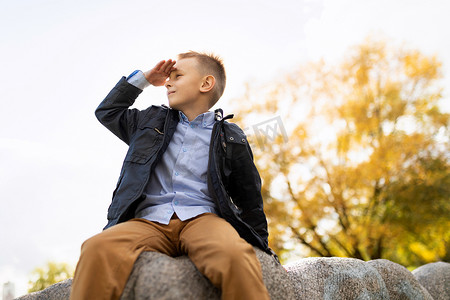 开朗淘气的男孩坐在石头上，手放在额头上眺望远方
