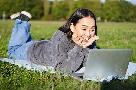 美丽女孩躺在公园里，惊讶地看着笔记本电脑、观看视频、在户外放松时与朋友聊天的肖像