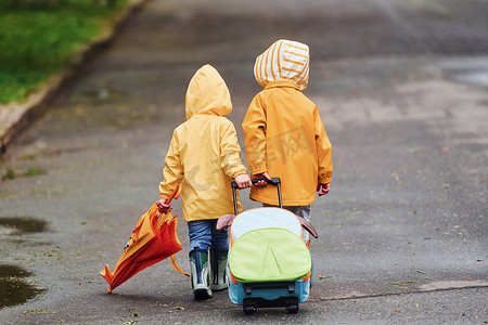 防水雨伞摄影照片_两个带着雨伞、手提箱和黄色防水斗篷和靴子的孩子在雨后一起在户外散步