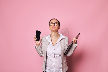 过度劳累的怀孕女商人站在粉红色背景的笔记本电脑和手机上。