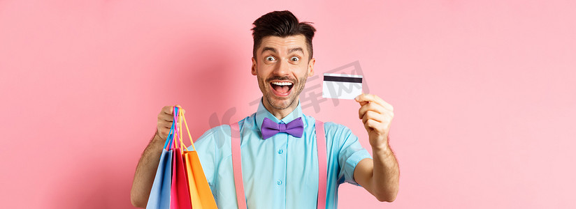 快乐的年轻人给信用卡，拿着购物袋，打折购物，站在粉红色背景中，微笑着