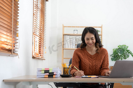 办公室的电脑摄影照片_在办公室工作的亚裔印度女董事坐在办公桌前，使用笔记本电脑分析商业统计数据，并持有图表，