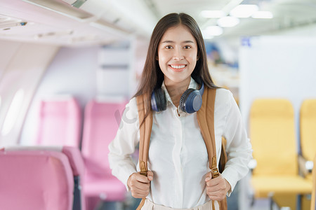 乘飞机旅行摄影照片_年轻的亚洲美女乘飞机旅行，乘客戴着耳机把手提行李放在飞机座位上方的储物柜里