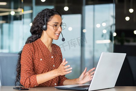 与同事进行视频通话在线会议，在现代办公室工作的西班牙裔女性，女商人微笑着使用笔记本电脑和耳机远程交谈