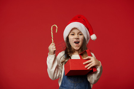 圣诞孩子们摄影照片_惊讶的小女孩戴着圣诞帽，拿着一根甜棒棒糖和红色礼盒，里面装着快乐的圣诞礼物