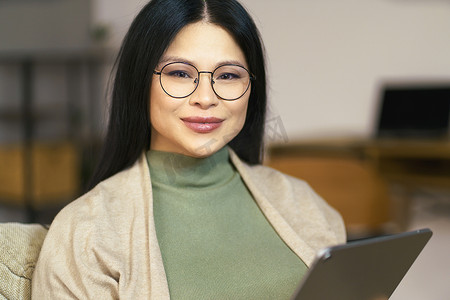戴着眼镜的快乐中年亚洲女性喜欢在平板电脑上浏览互联网，同时舒适地坐在家里