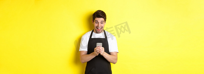 咖啡师在手机上阅读信息时看起来很惊讶，站在黄色背景的黑色围裙里