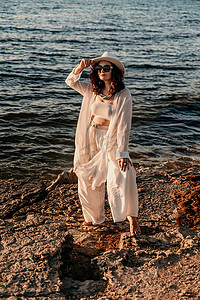 男式裤装摄影照片_一位穿着白色裤装和帽子的女人站在沙滩上享受大海。