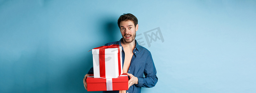 蓝色礼物摄影照片_留着胡须的帅哥拿着情人节礼物送给情人，拿着礼物站在盒子里，看着相机，蓝色背景