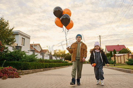 万圣节，可爱的孩子、男孩和女孩、带着橙色黑色气球的兄弟姐妹沿着乡村街道走