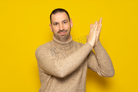 40 多岁留着胡子的西班牙裔男子穿着米色高领毛衣，满意地拍着手，双手放在一边，在黄色背景中被隔离。