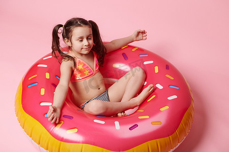 可爱的小女孩在粉红色充气游泳环甜甜圈上以荷花的姿势沉思，粉红色背景