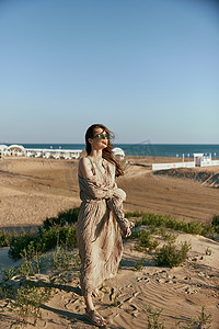 在远处可见的大海背景下，一位身穿米色连衣裙的优雅女性在沙滩上行走