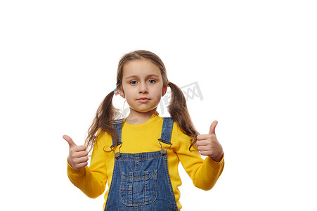 蓝色小学摄影照片_留着两条马尾辫、穿着黄色毛衣和蓝色牛仔工装裤的可爱女婴，看着镜头竖起大拇指