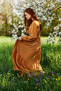 一个快乐、苗条、甜美的女人穿着一件橙色的长裙站在一棵开花的树附近的高高的草丛中，她开心地微笑着掀起裙子的下摆，望向别处