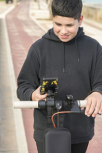14-17 岁的前视图在自行车道上快乐地微笑着骑电动滑板车，并使用智能手机音乐应用程序，E-Mobility 城市休闲概念