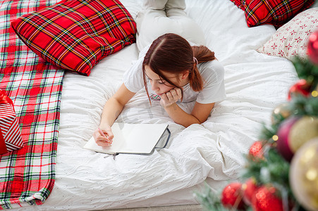 快乐的少女用笔和笔记本在圣诞树上躺在床上做愿望清单或做新年清单。