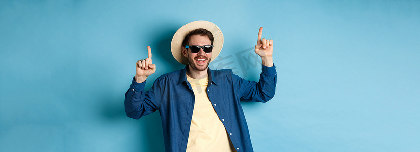 戴着草帽和太阳镜的快乐的白种人，站在蓝色背景上，在假期里跳舞和玩乐。