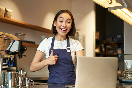 亚洲女孩围裙的肖像，咖啡店的咖啡师，竖起大拇指，赞成或推荐 smth，在咖啡馆工作，笑得开心
