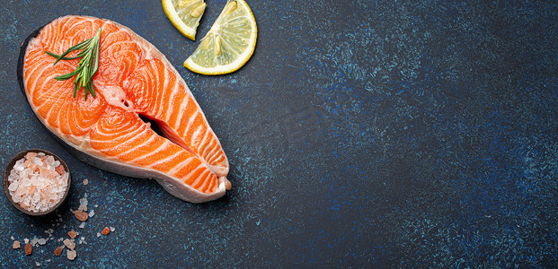 深蓝色质朴石背景上带迷迭香的新鲜生鲑鱼片牛排，玫瑰盐和柠檬顶视图，健康均衡的饮食和营养
