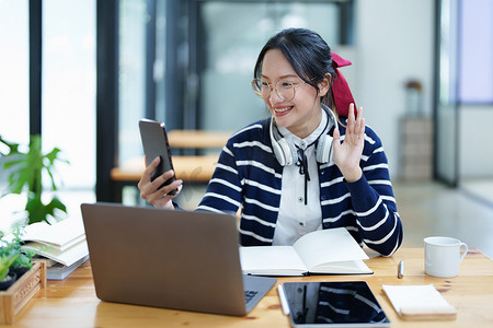 一位亚洲少女使用智能手机、戴着耳机并使用笔记本在图书馆的木桌上通过视频会议在线学习的肖像