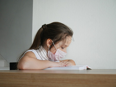 在 Covid-19 大流行期间，一个女孩脸上戴着医用口罩在家看书并做学校作业。