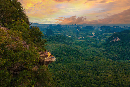 山龙虎山摄影照片_泰国甲米龙冠山，一位年轻的旅行者坐在日落时悬在深渊上的岩石上