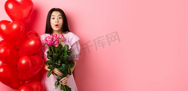 穿着裙子的亚洲女孩惊讶地站在情人节心形气球附近，对着镜头说哇，手捧情人送来的花束，浪漫的玫瑰约会，粉红色背景