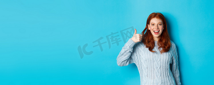 穿着毛衣的快乐红发女孩，站在蓝色背景上，竖起大拇指表示赞同，喜欢并赞美产品