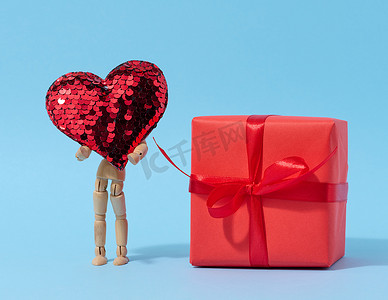 一个木制人体模型拿着一颗由亮片制成的红心和一个用红纸包裹的盒子，祝贺假期
