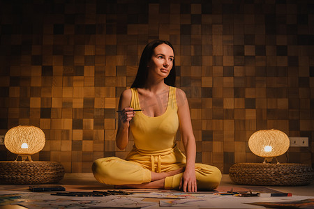 一个穿着黄色衣服的女孩坐在莲花位置上，在家里用晚上的灯光在纸上画画
