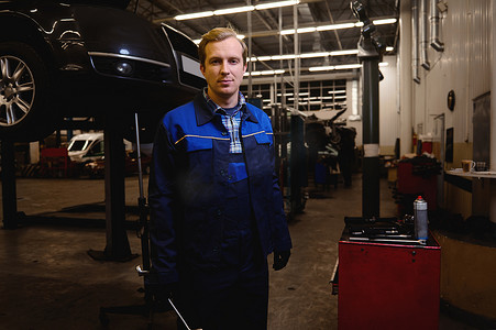 一名技术员、汽车机械师、汽车工程师站在一辆举起的汽车的背景下，手里拿着扳手，在维修店或汽车维修车库工作