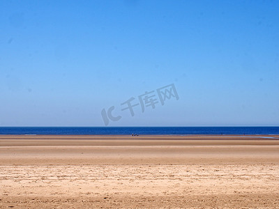 一个长长的沙滩，有蓝色的夏日天空和蓝色的大海，在水边有一群遥远的无法辨认的人