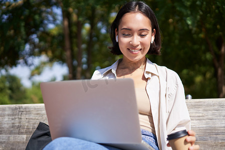 亚洲女孩坐在笔记本电脑和无线耳机旁，喝咖啡，看屏幕，做作业，远程工作